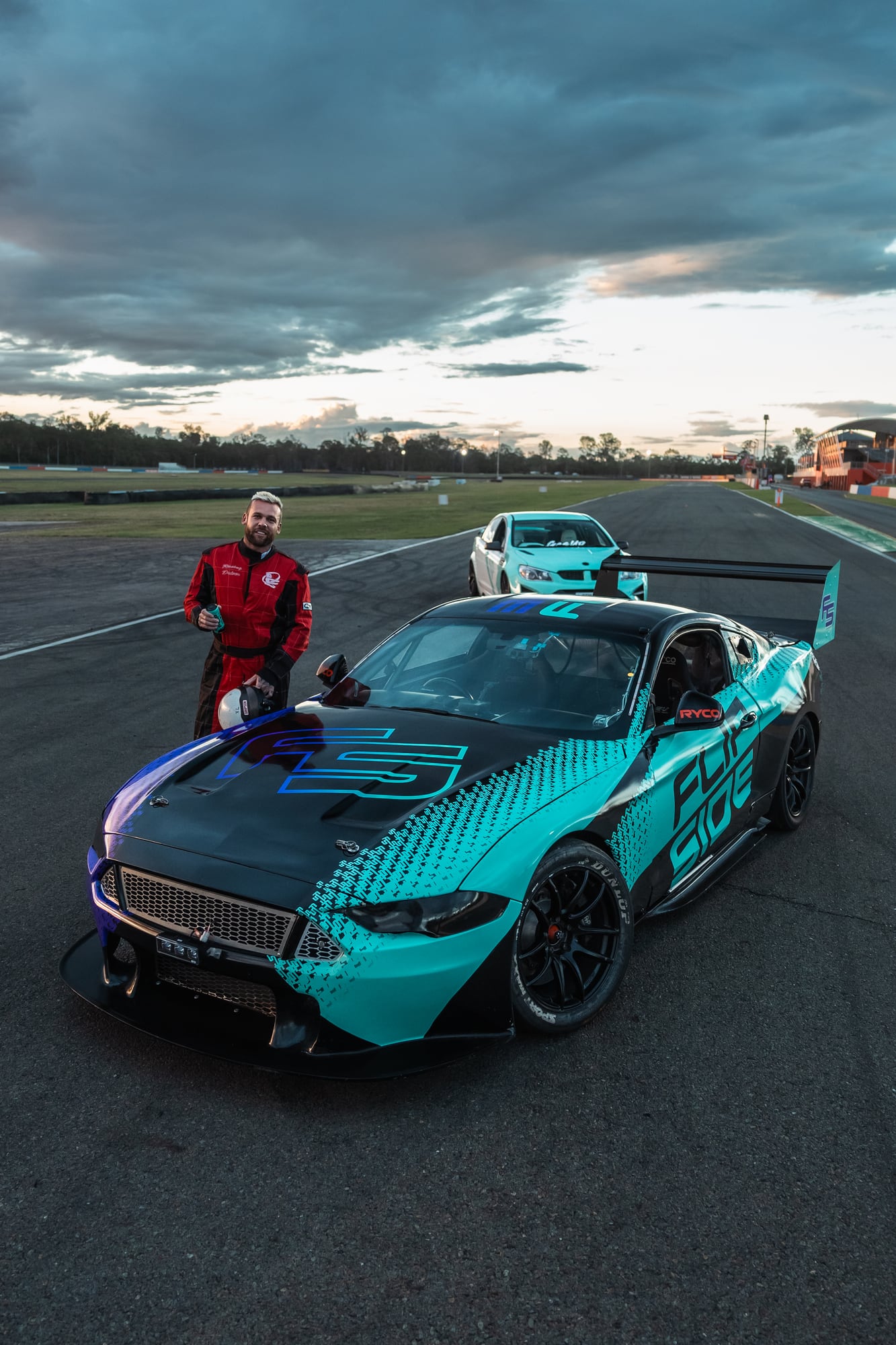 A Race X race car on the pit straight at dusk, its driver standing alongside.